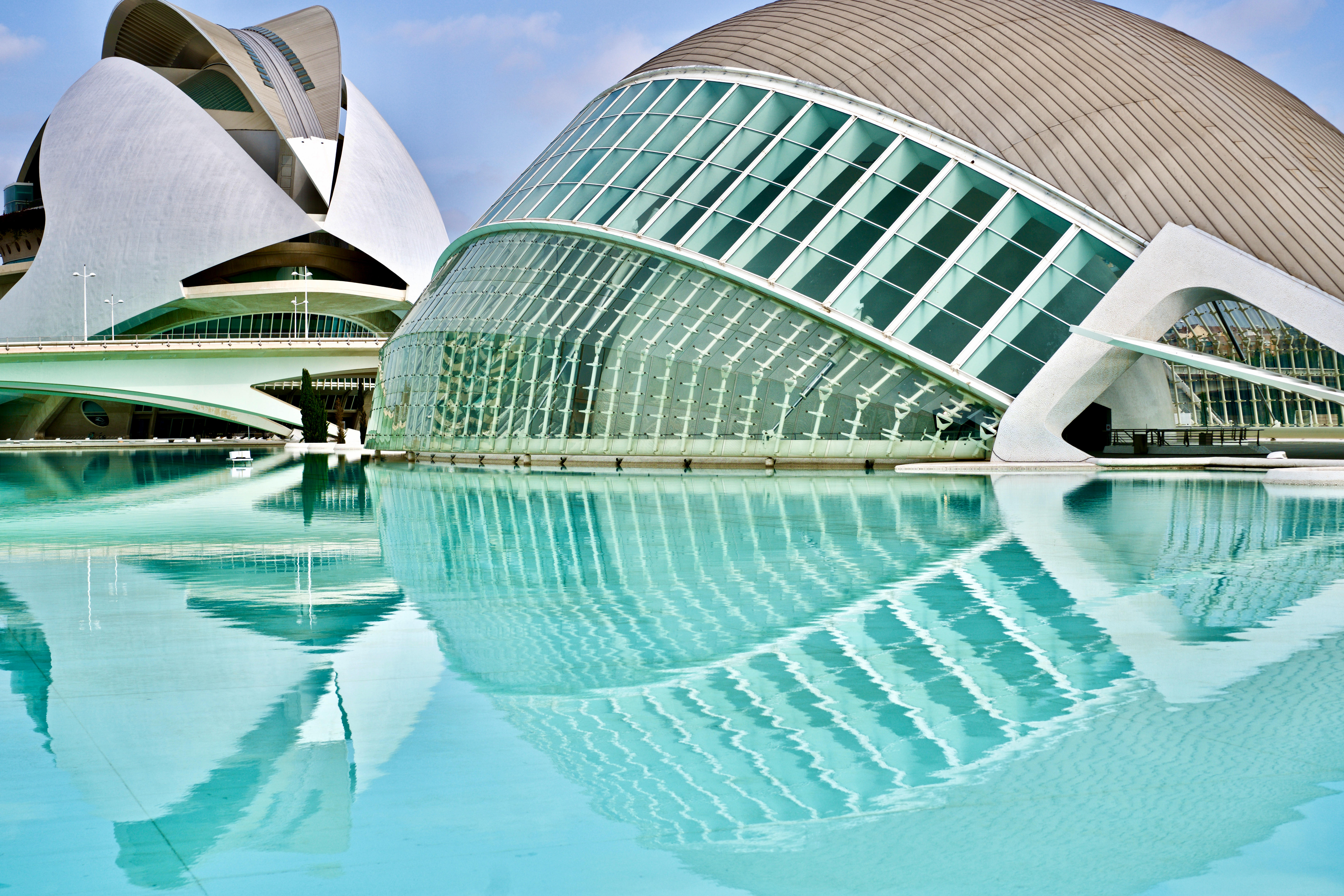 Reflection of Calatrava's amazing Opera House in Valencia, Spain. Two modern, futuristic buildings with large, curved glass and metallic surfaces are reflected in a serene body of turquoise water.