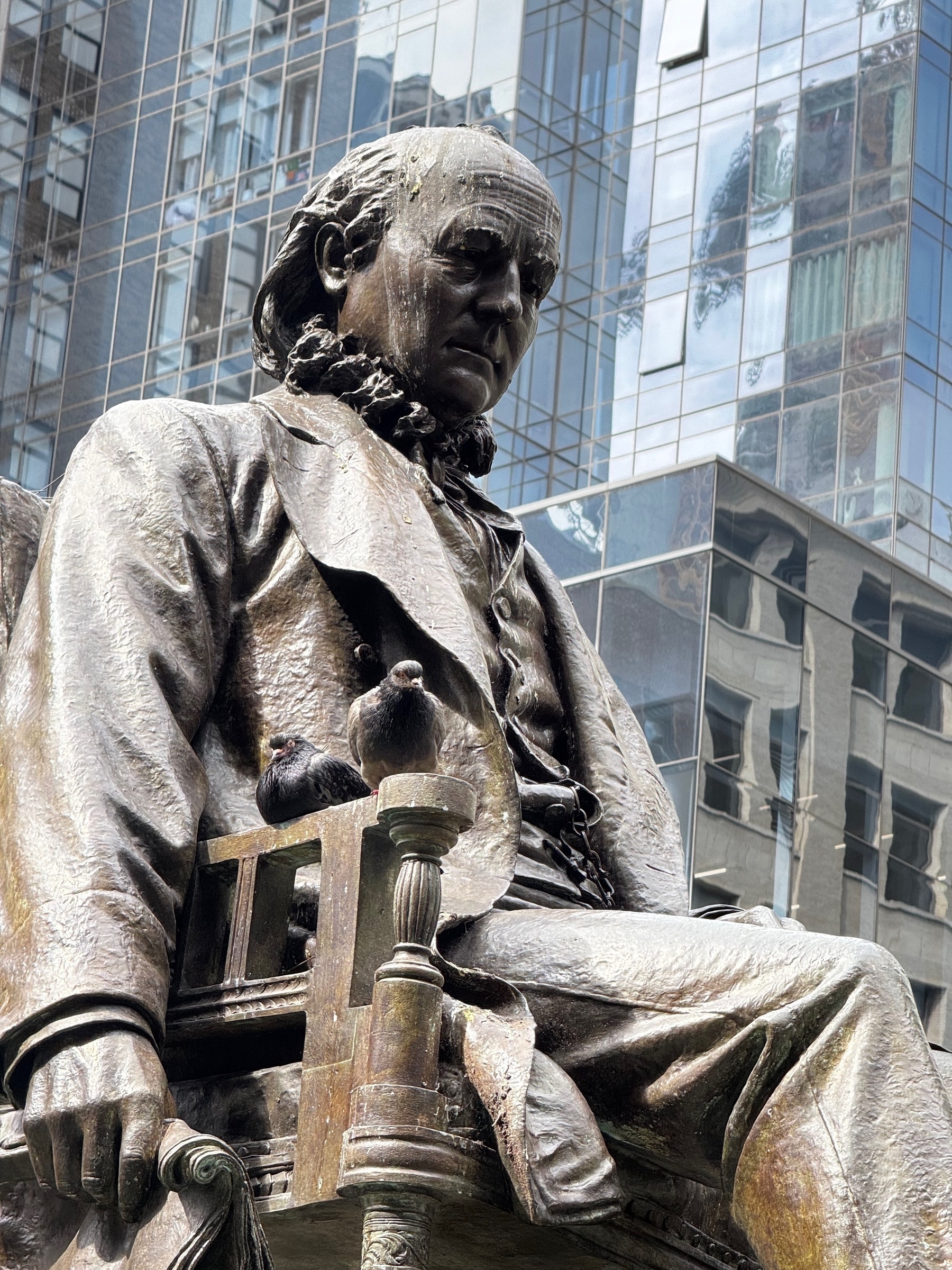A bronze statue of a seated man, Horace Greely, with pigeons perched on it is set against a backdrop of modern glass buildings.