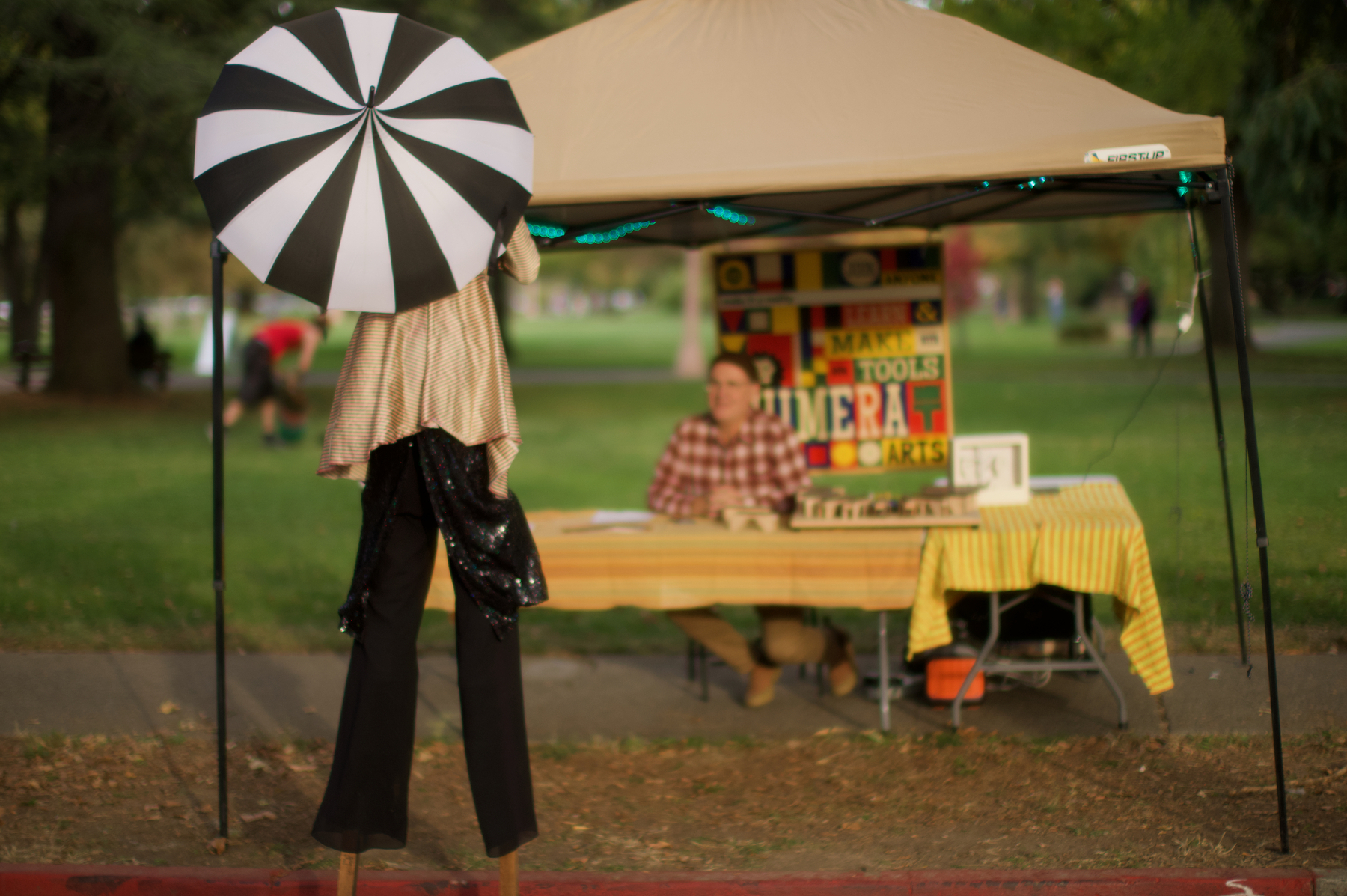 A person in a costume stands on stilts holding a black-and-white striped umbrella in front of a booth with a seated man and various items displayed on a table.